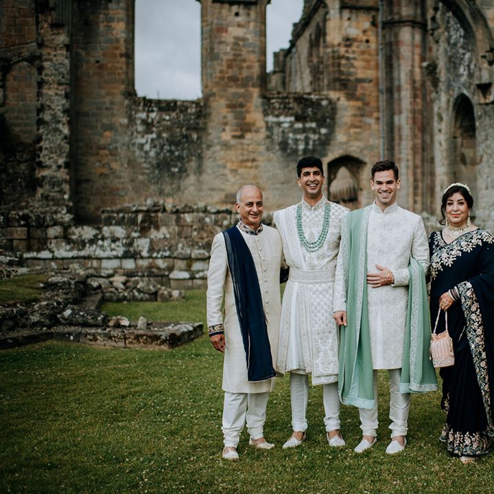 Grooms wear white and sage green sherwani's as they stand outdoors with their family 