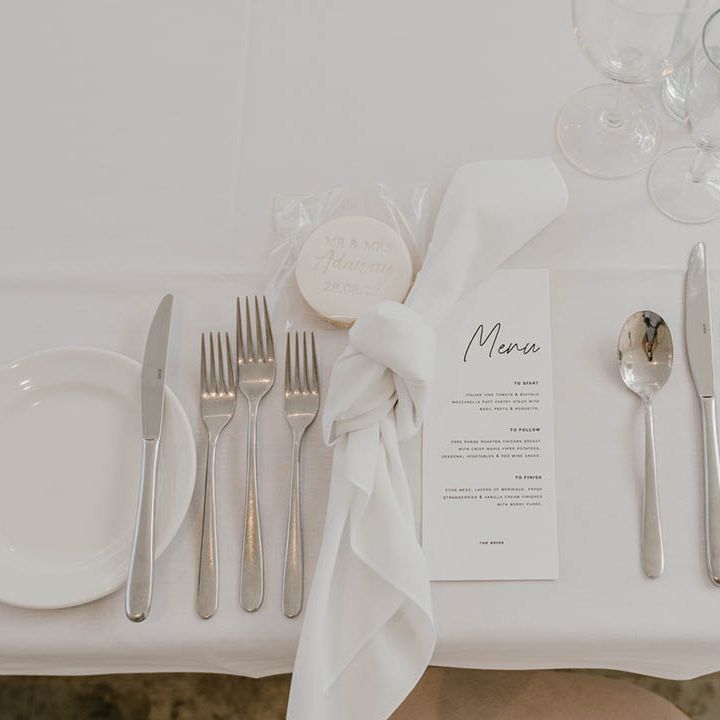 All white wedding place setting with black and white wedding menu with silver cutlery and hand made biscuit wedding favours and white wedding napkin