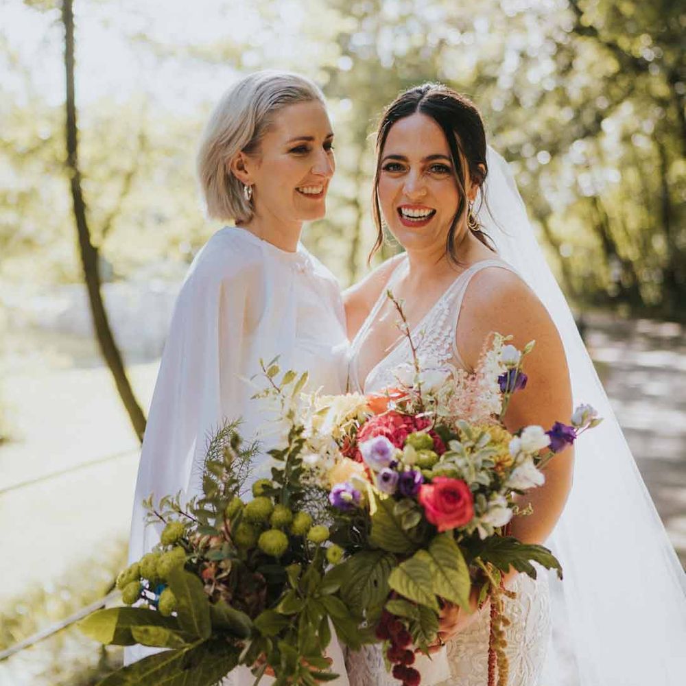 Bride in v neck wedding dress and church length veil standing with bride in bridal jumpsuit with cape both holding large wildflower and colourful floral bouquets - wedding budget spreadsheet 