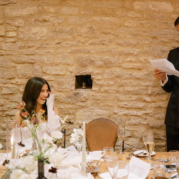 Groom in dark double breasted navy suit standing up reading out his groom speech with the bride sitting down enjoying the speech at the reception 