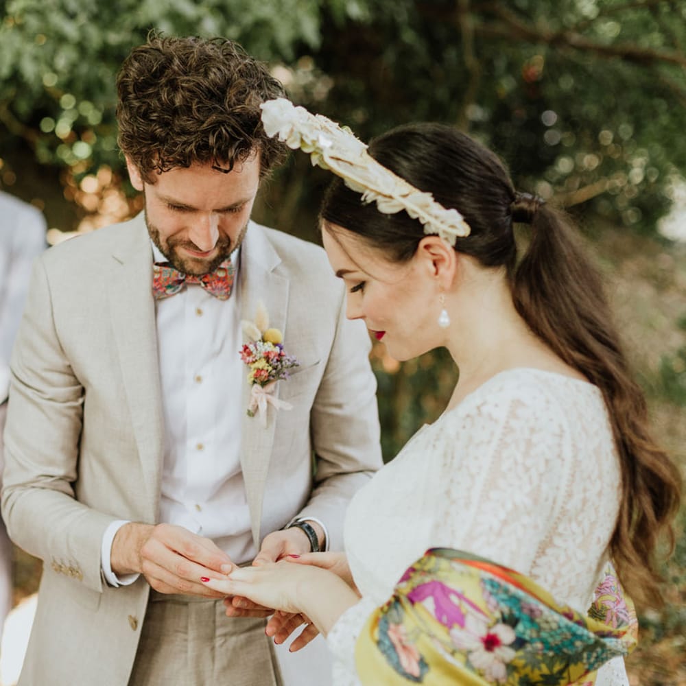 Bride in white lace wedding dress and boho bridal vrown exchanges rings with groom in greige suit and colourful bow tie