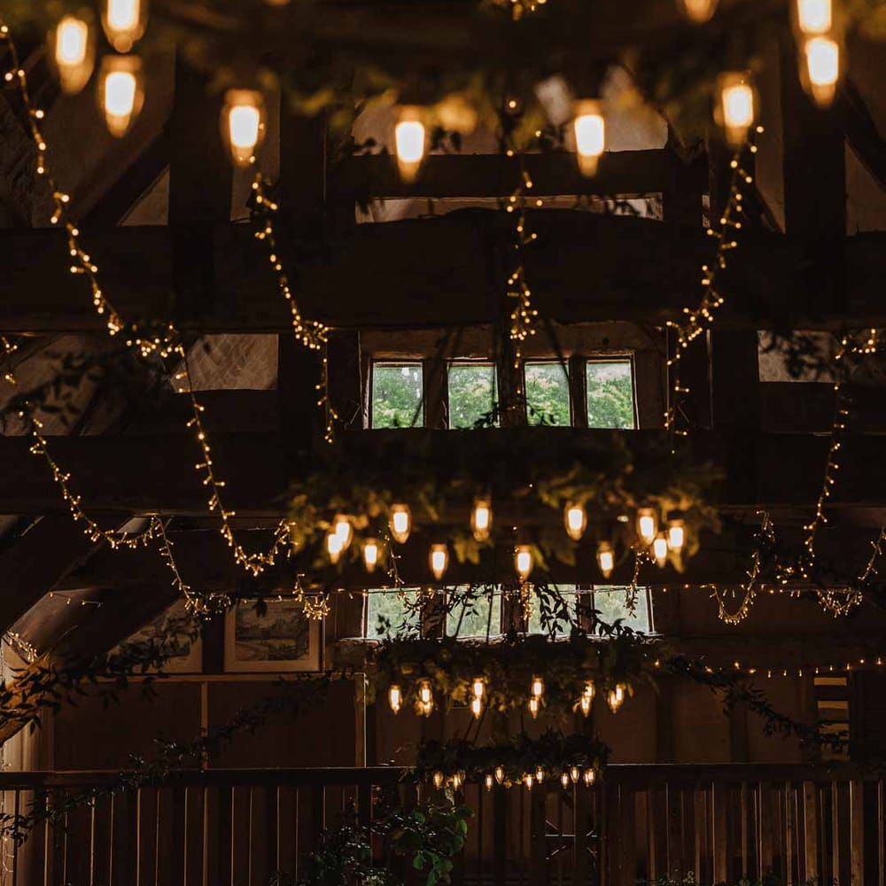 Bride and groom standing in Lains Barn reception room with fairy lights, suspended foliage arrangements, festoon chandeliers, large neon wedding sign, foliage wall and candles 