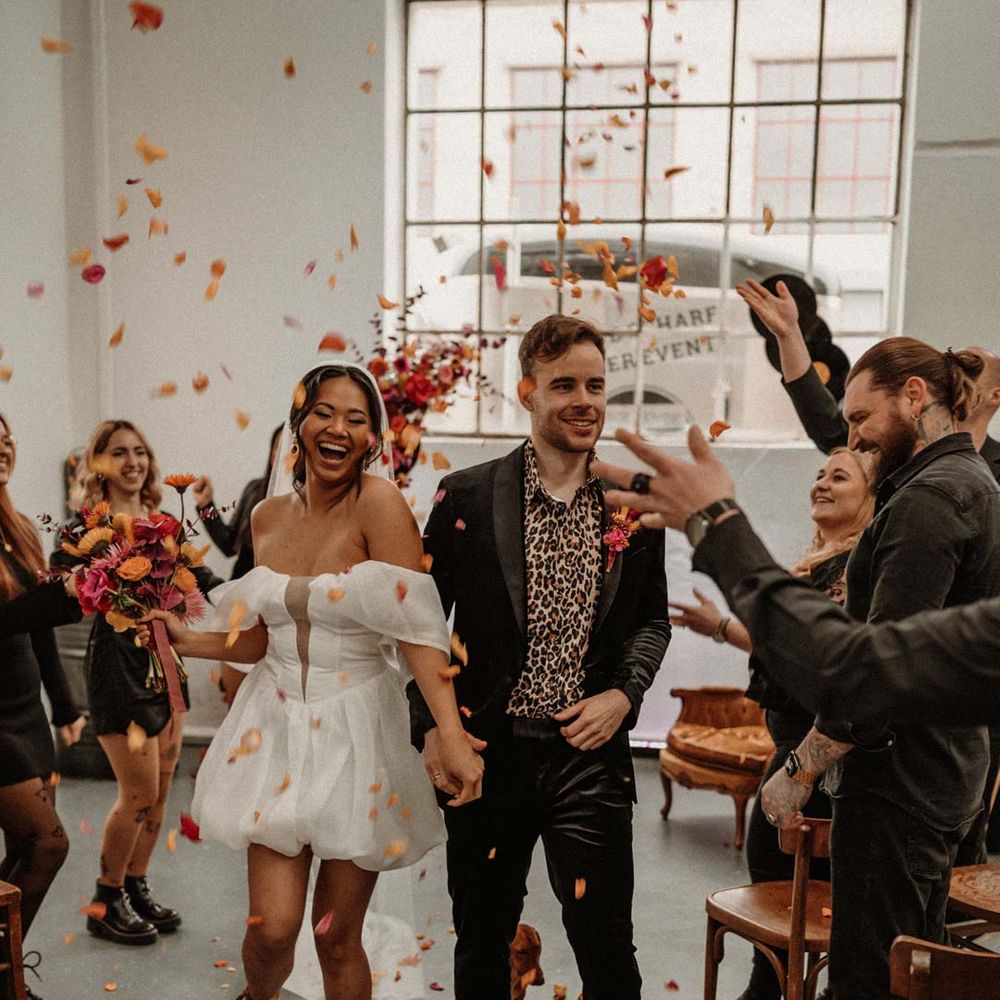 Groom in wet look black grooms suit with leopard print shirt and pink and orange boutonniere standing by vinyl wedding arch walking hand in hand with bride doing colourful wedding confetti exit with bride in off shoulder tulle short wedding dress with church length veil and white platform Dr Martens wedding shoes