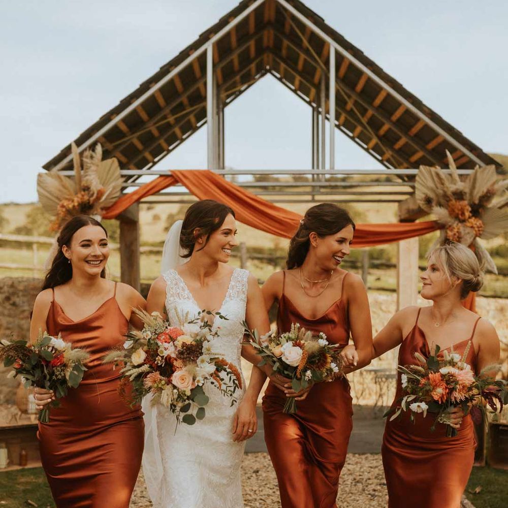 Bridesmaids in burnt orange satin midi cowl neck bridesmaid dresses walking with bride in lace wedding dress 