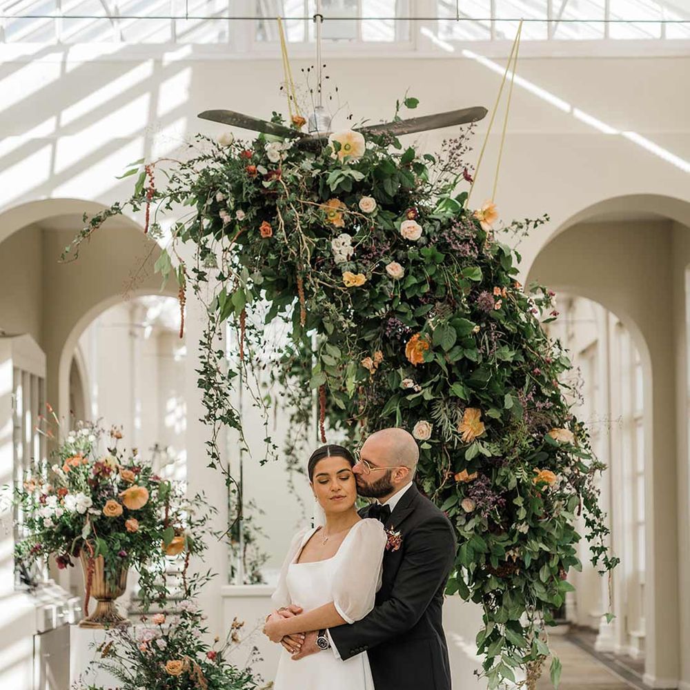 Groom in classic black tuxedo with bowtie and wildflower boutonniere dancing with bride in short sheer puff sleeve wedding dress with square neck and wrist loop with sheer pearl embellished bridal veil surrounded by suspended wildflower and foliage arrangements in large gold vases at Buxted Park
