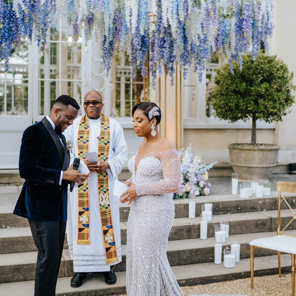 Bride in illusion v neck sheer off shoulder long sleeve pearl embellished wedding gown standing with groom in groom in velvet midnight blue double breasted groom’s blazer, velvet bowtie and peach garden rose boutonniere underneath suspended pastel flower arrangements at Syon Park vow renewal 