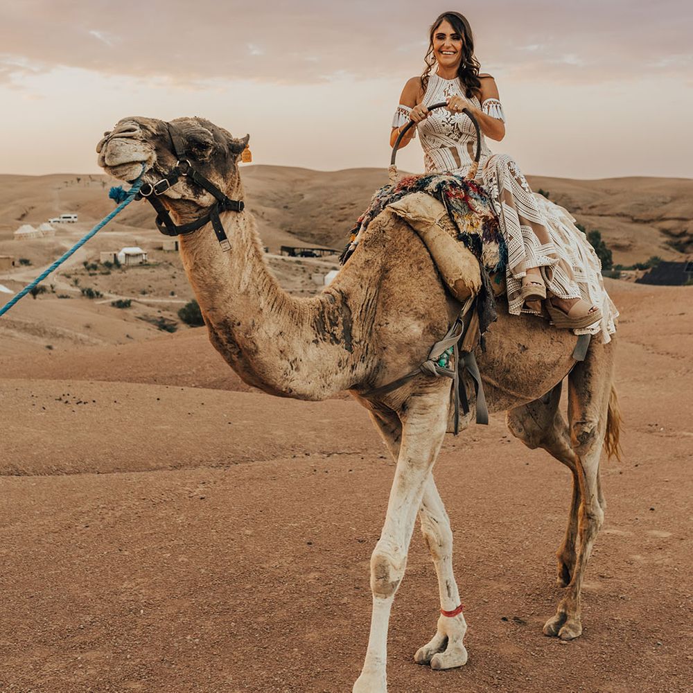 The bride in a boho lace tassel wedding dress rides upon a camel at destination wedding 