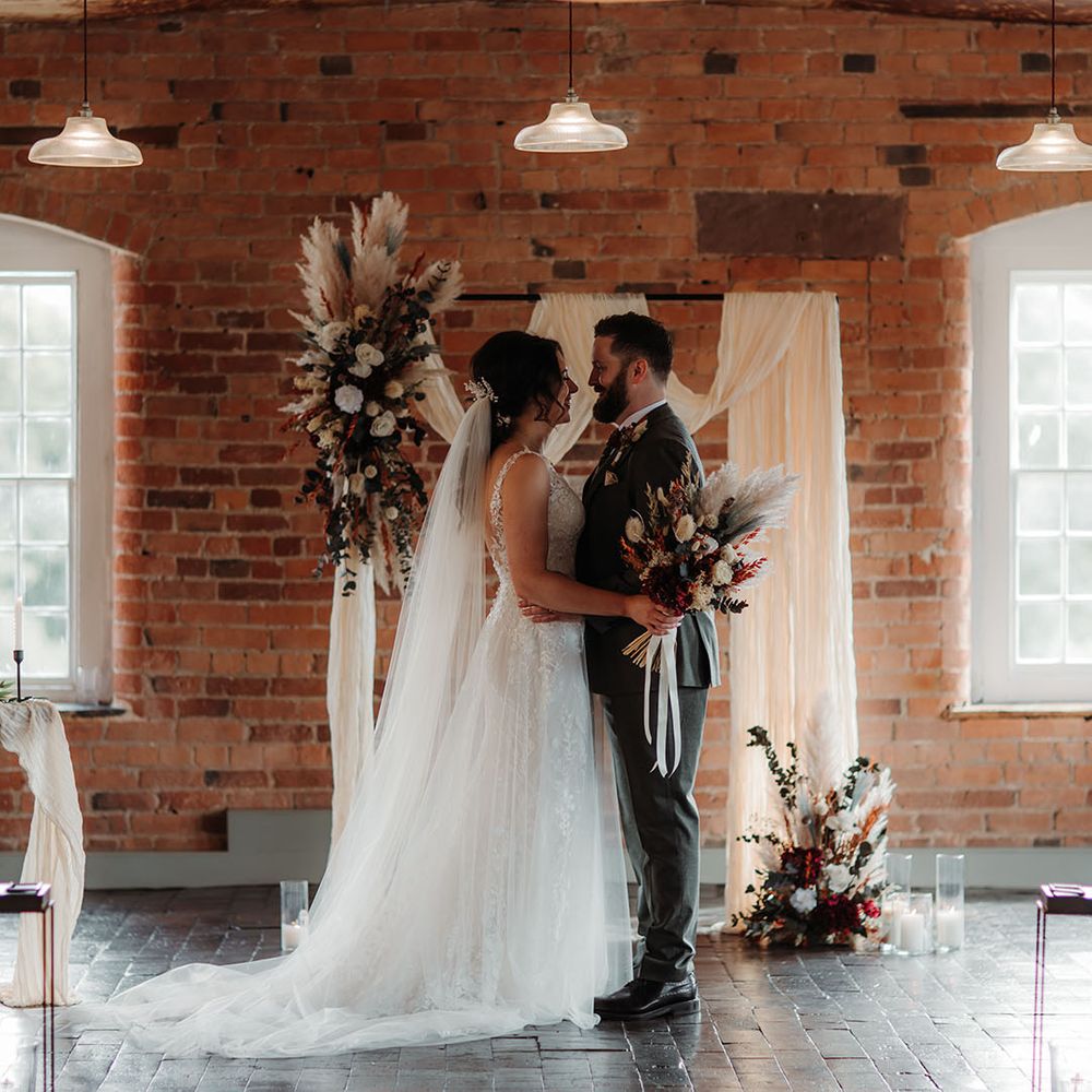 The bride and groom stand in front of the boho altar decoration made from white drapery and dried flower arrangements on wooden frame 