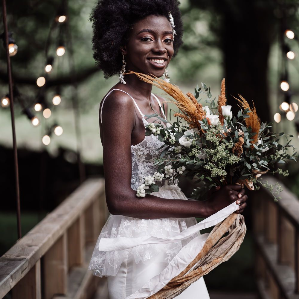 Black bride with afro hair wearing a lace peplum wedding dress holding a orange flower and foliage wedding bouquet tied with ribbon