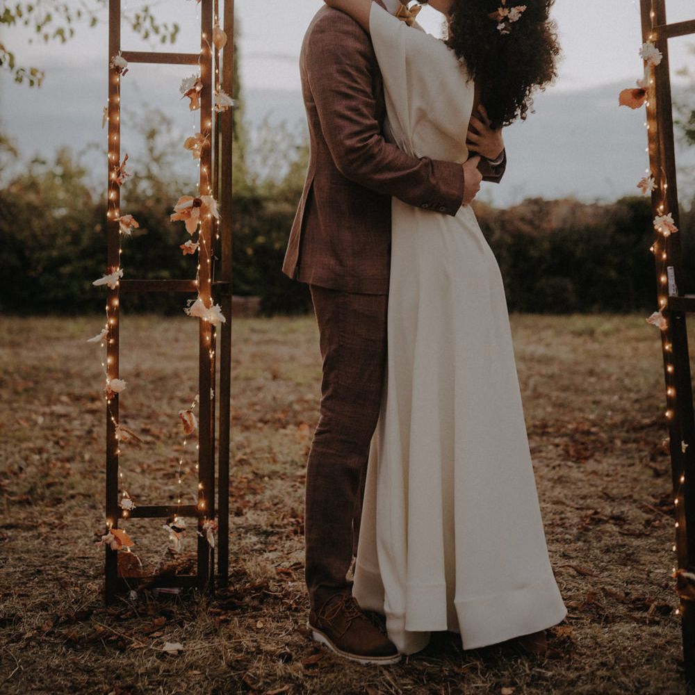 Groom in a brown suit kissing his bride with naturally curly hair under a fairy light decorated pergola 