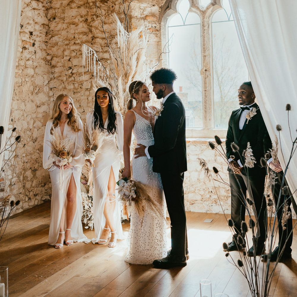 Bride in a appliqué wedding dress with pearl headband standing at the alar with her groom in a tuxedo and bridesmaids in satin dresses 