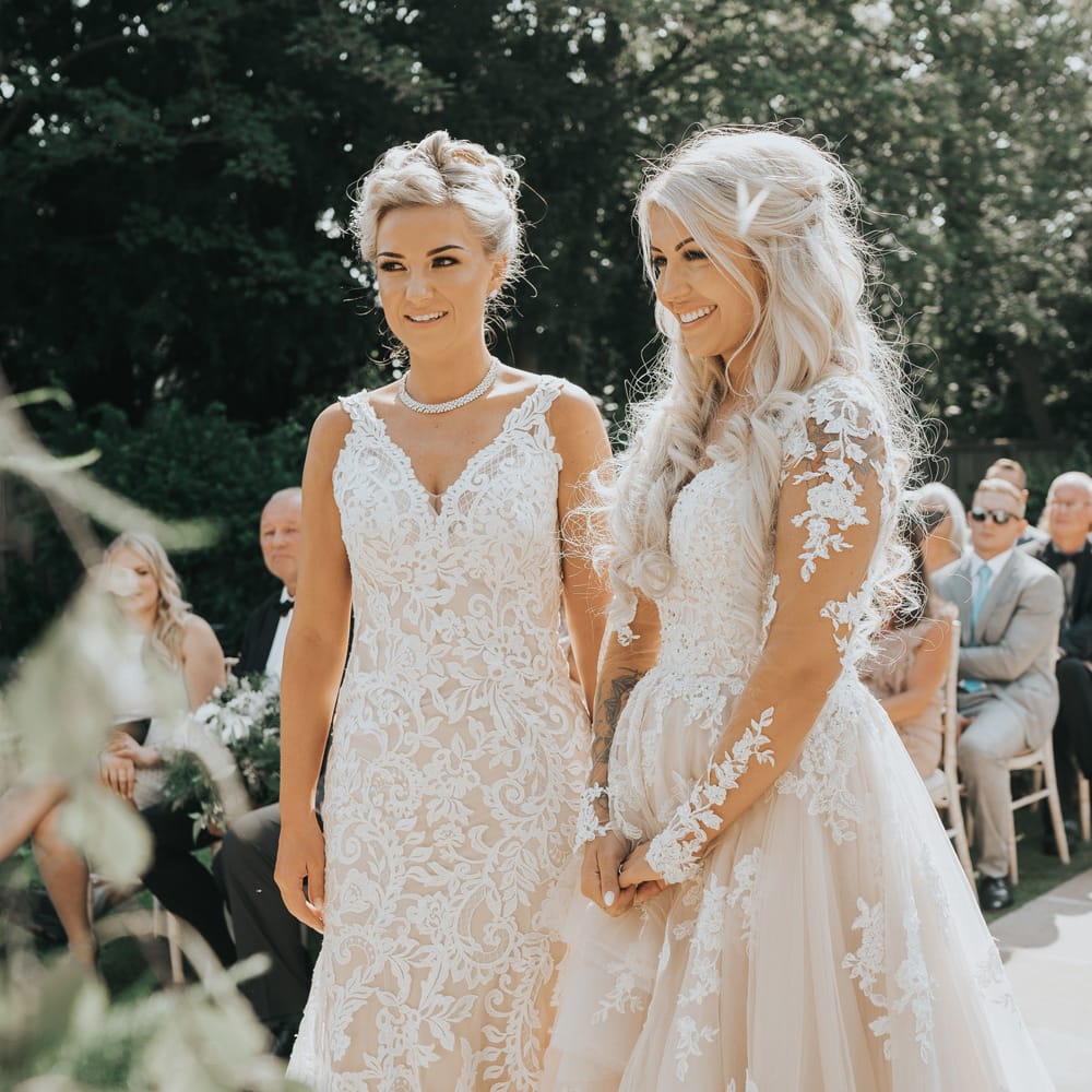 A lesbian couple stand at the alter during their wedding ceremony. They are both blonde. One wears her hair up and the others is long and curly in a half up half down style. 
