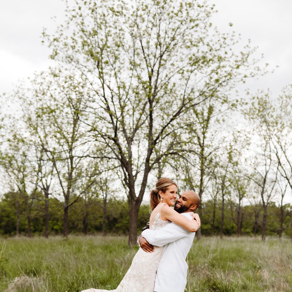 Groom in white tuxedo jacket lifts up bride in white embroidered Made With Love wedding dress in field at backyard wedding