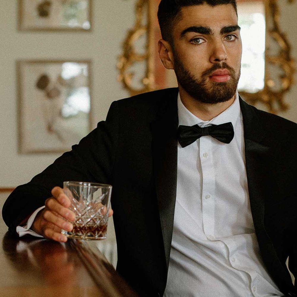Groom in a tuxedo drinking whisky at the bar at Bourton Hall wedding venue 