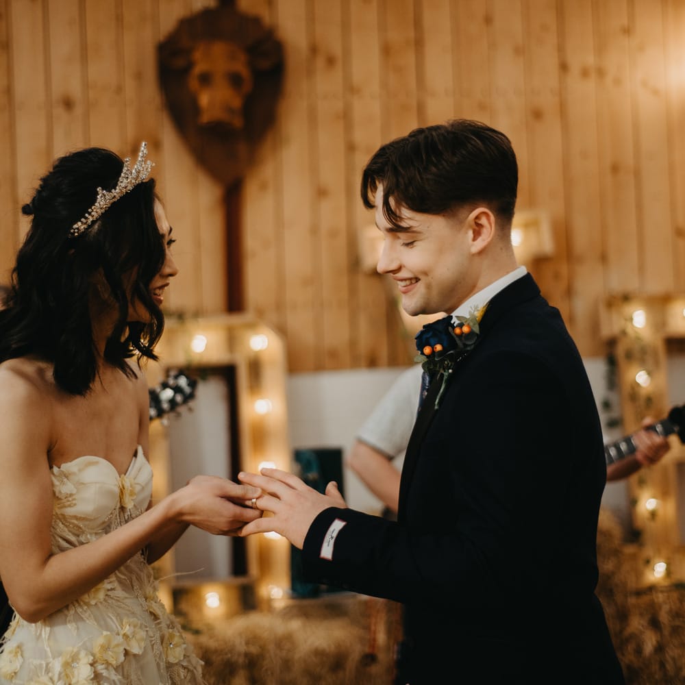 Bride in strapless feathered wedding dress and headpiece puts rings on grooms finger as he wears blue suit and floral buttonhole at rustic wedding in Lancashire