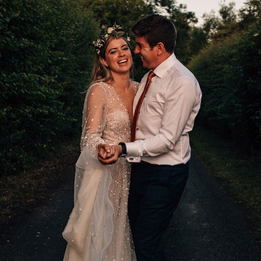 Bride in boho flower crown and long sleeve Julie Vino wedding dress smiles whilst hugging groom in white shirt during golden hour