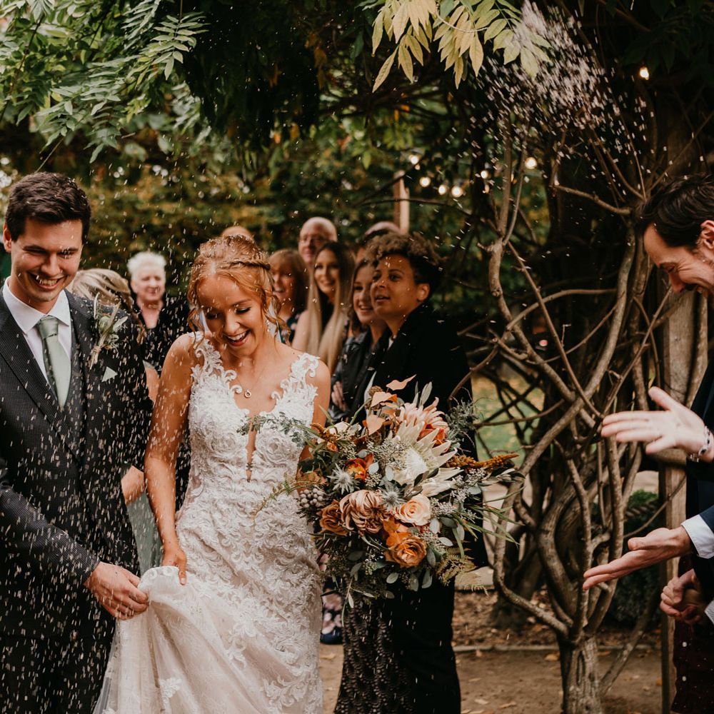 Groom in dark suit and light blue tie holds tulle skirt of brides wedding dress as guests throw multicoloured confetti after rustic wedding ceremony in barn