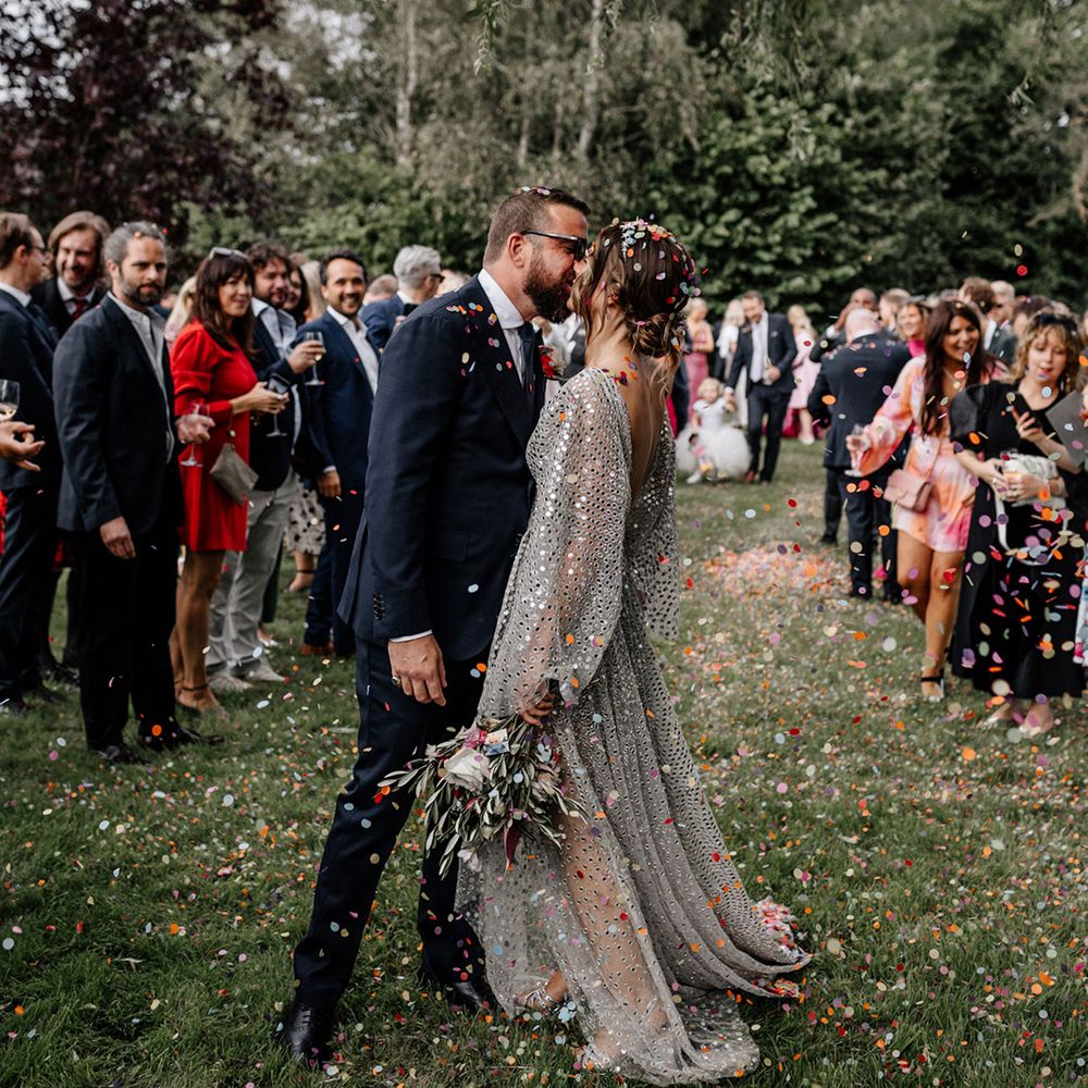 Bride in a silver sequin wedding dress and groom in a navy suit kissing at the end of the confetti line 
