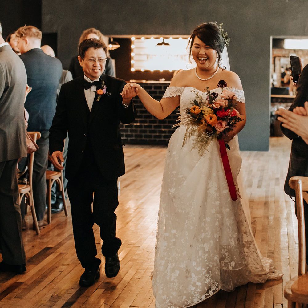 Bride in lace off the shoulder wedding dress and pearl necklace holding pink and red bridal bouquet walks down the aisle holding hands with father of the bride in black tie and colourful floral buttonhole