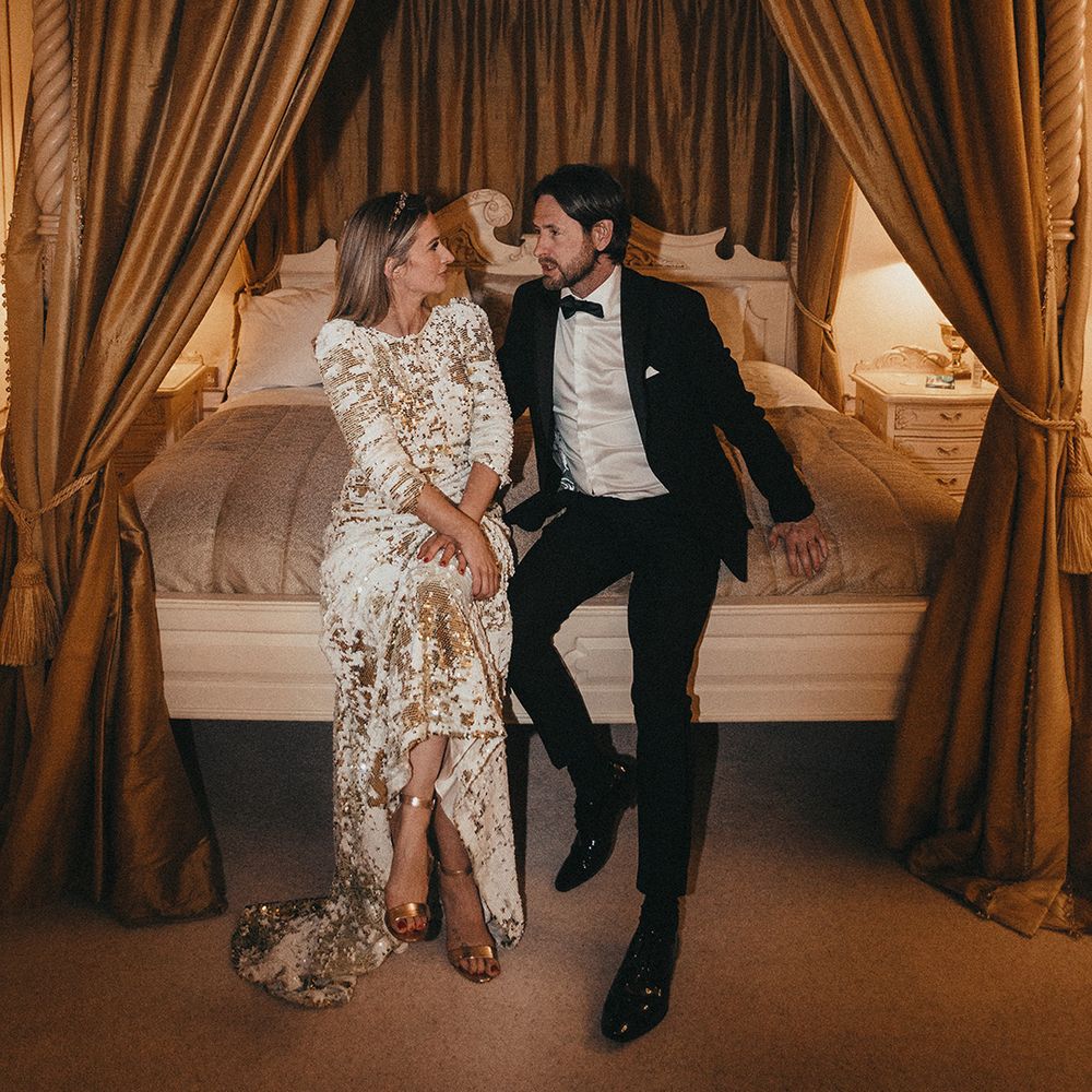 Bride & groom sit at the front of four poster bed as bride wears Abigail Of Gardenia sequin gown and groom wears black tie 