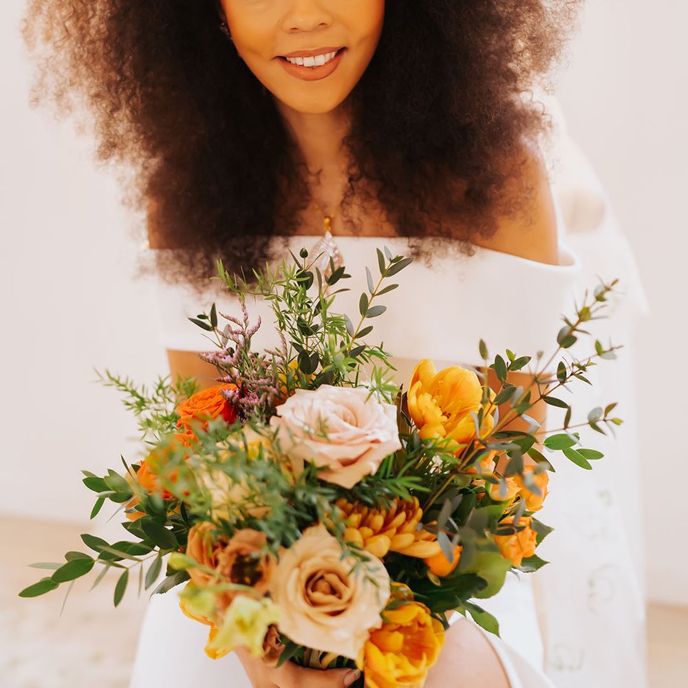 Black bride with afro hair wearing a hair accessory and holding an orange, yellow and white golden wedding bouquet 