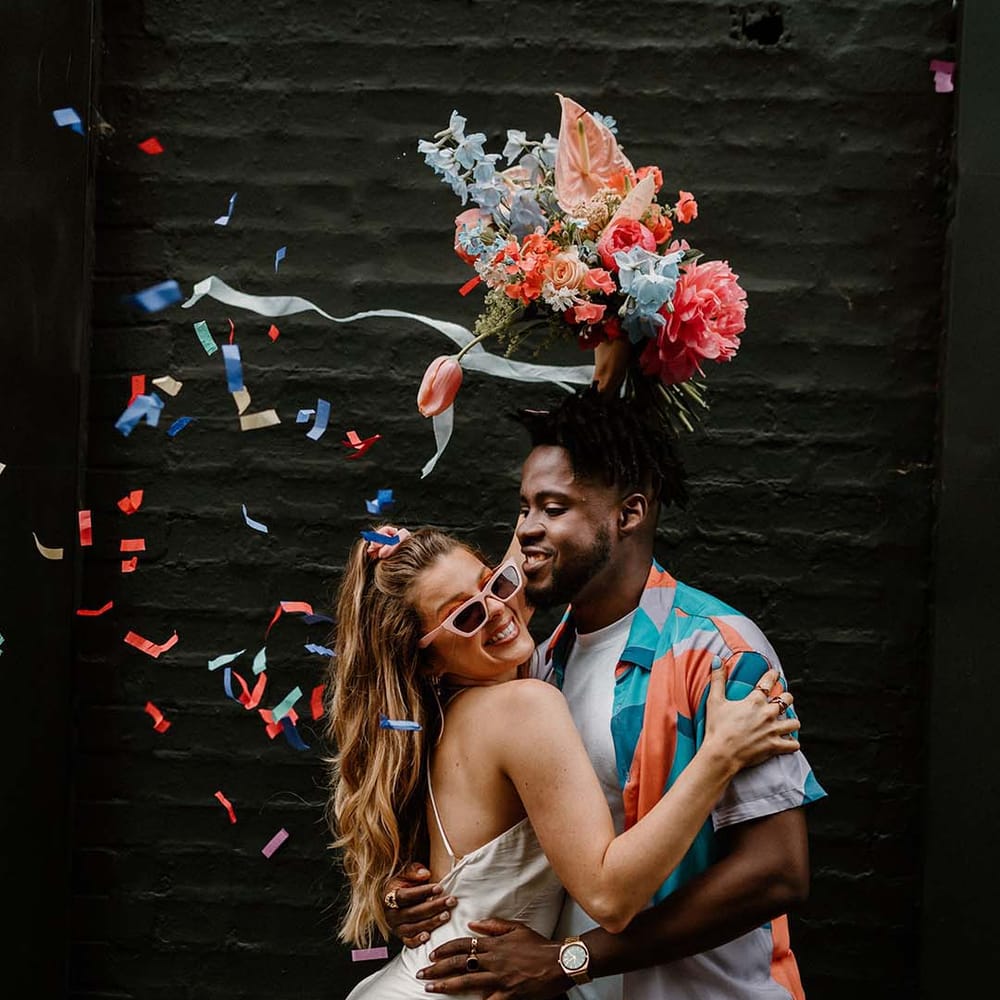 Colourful confetti moment at city elopement with bride in a short wedding dress holding a pink and blue bouquet in the air and groom in a patterned shirt and white tee