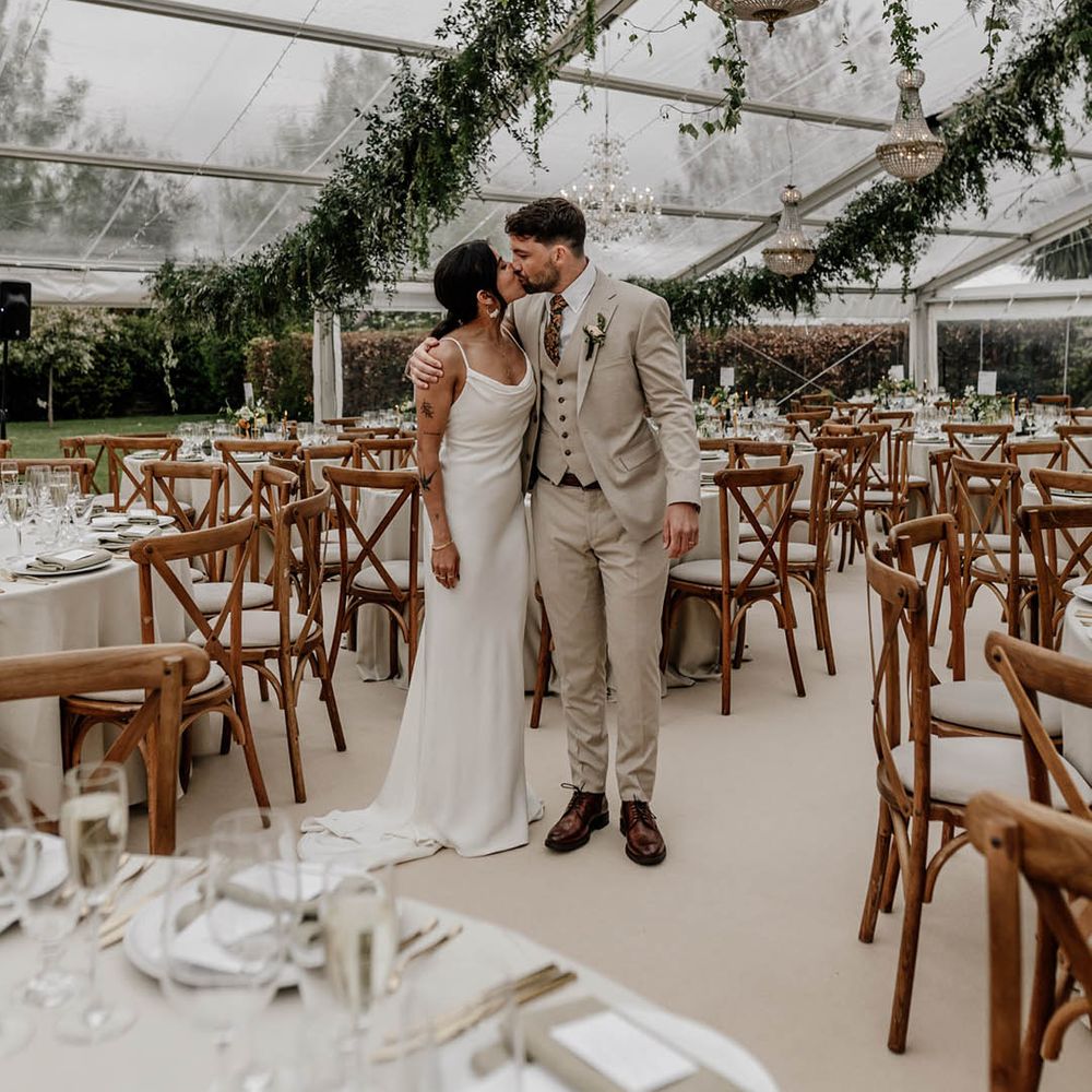 Bride in a satin wedding dress and groom in a beige suit kissing in the middle of their clear glass marquee wedding reception with wooden furniture, greenery decor and handing chandelier 