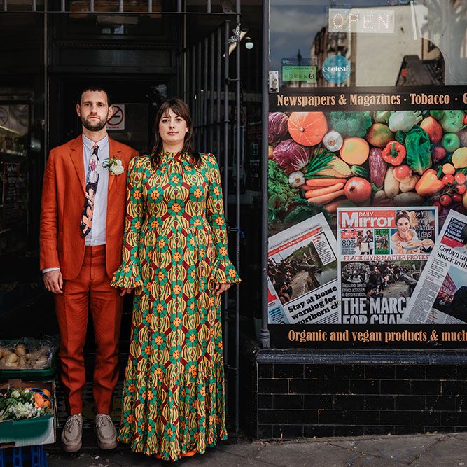 Bride & groom stand in shop door on their wedding day in Bristol as bride wears brightly coloured dress and orange suit 