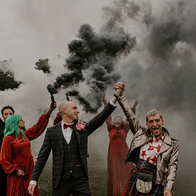 Grooms walk through grey smoke bombs held by their bridesmaids in deep red dresses 