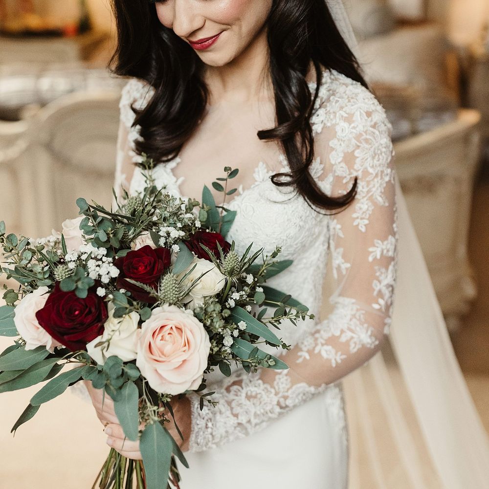 Bride wears red lipstick and her dark hair in loose curls complete with pearl hair accessory whilst holding white and red floral bouquet 