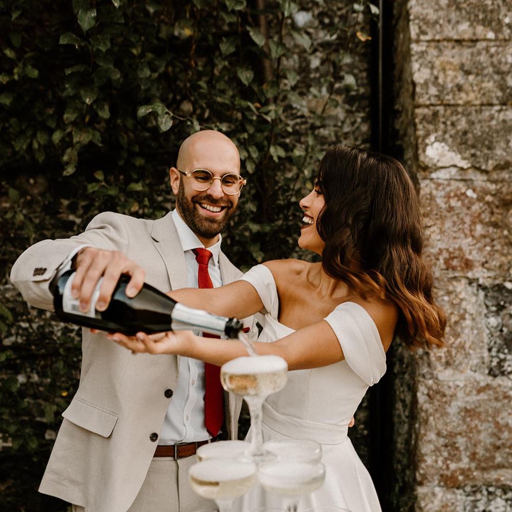 Bride and groom smile brightly as they pour champagne onto their champagne tower 