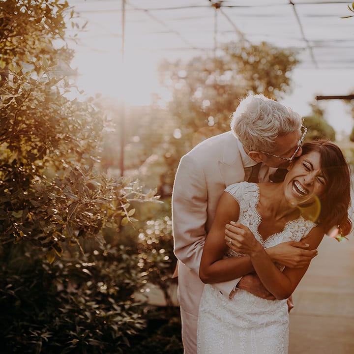 Groom hugs Bride from behind and kisses her on forehead while she laughs with greenery in the background