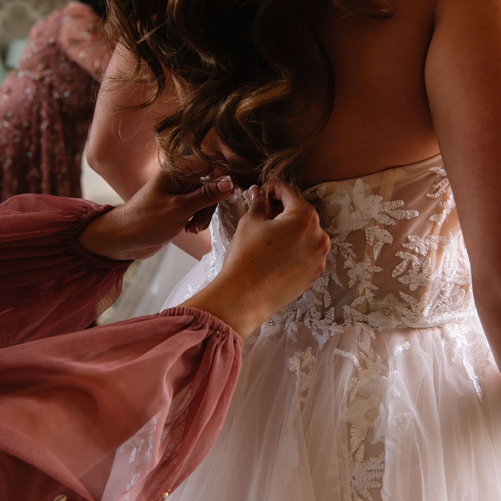 Bride with curled brown hair being buttoned into her strapless lace wedding dress with flowers 