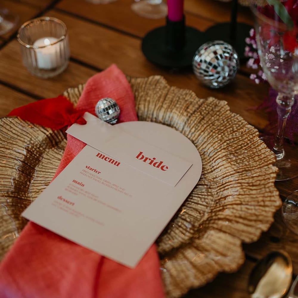 Round gold charger plate with rounded retro wedding menu, bright pink napkin, gold cutlery and wedding disco ball decorations
