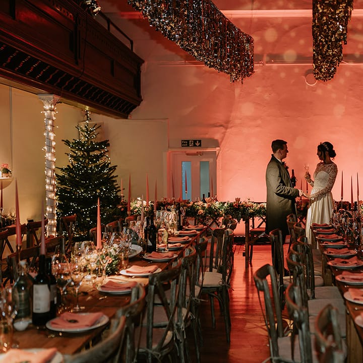 Bride and groom sharing a glass of wine in the reception room of London wedding venue with gold wedding streamers, projections, orange tapered candles, orange napkins, orange rose and foliage tablerunners and fairy lights