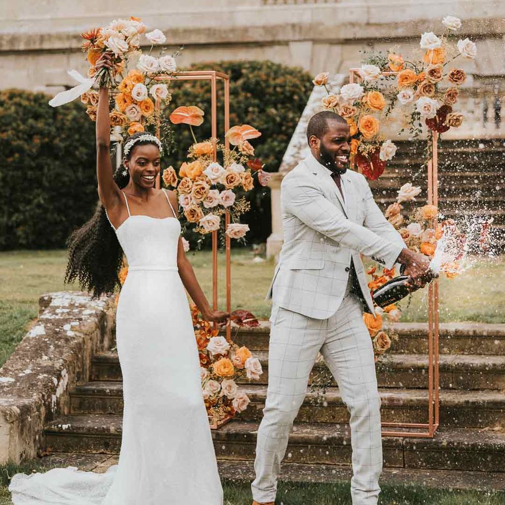 Bride in strappy satin wedding dress and groom in light grey suit doing champagne pop wedding photo at Kirtlington Park wedding venue 