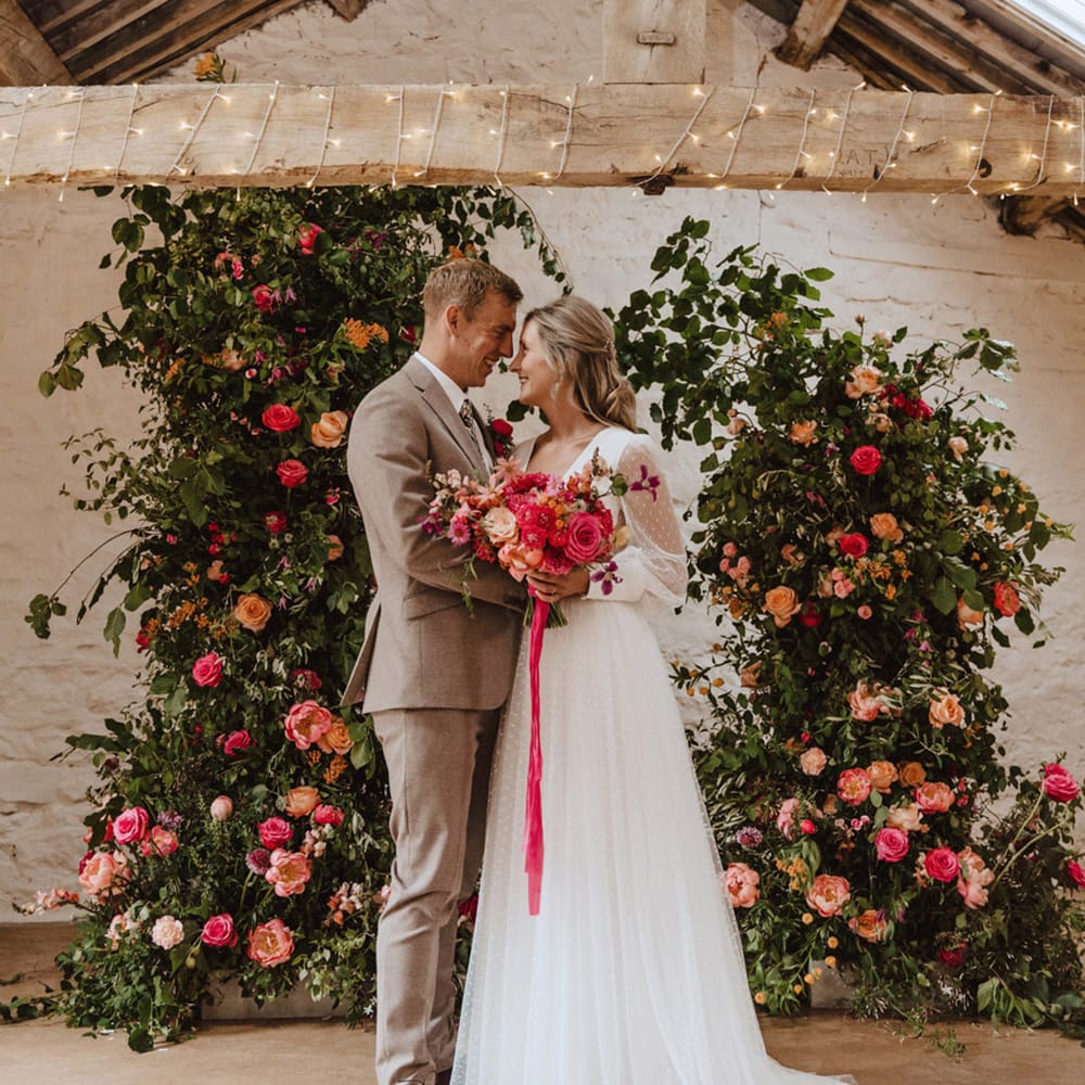 The bride wears a Sassi Holford wedding dress with long sheer polka dot sleeves with the groom in a beige suit in front of the pink wedding column flower arrangements 