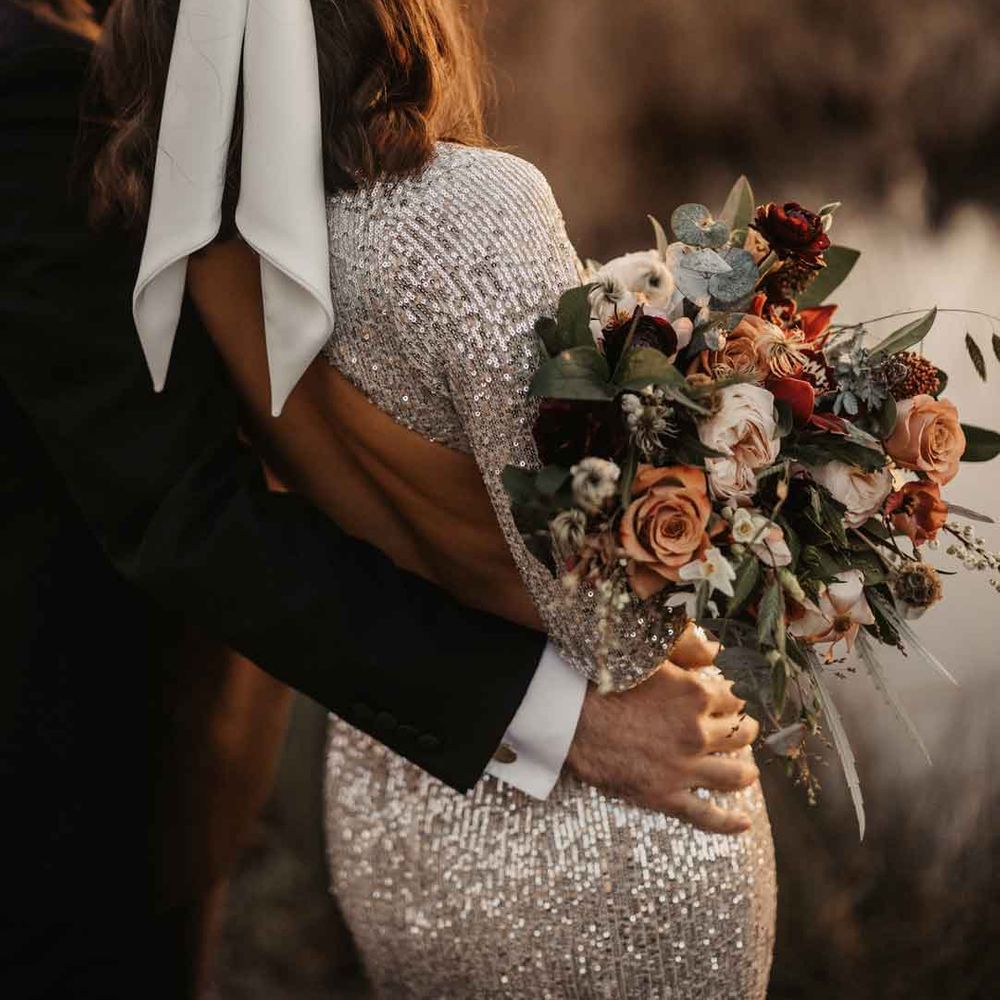 Bride in silver sparkly wedding dress with open back detail and large white satin bow hair bridal hair accessory with groom in classic black tuxedo looking out at the lake holding bouquet with garden roses, carnations, peonies, eucalyptus, baby's breath and dried flowers tied with mustard velvet ribbon 