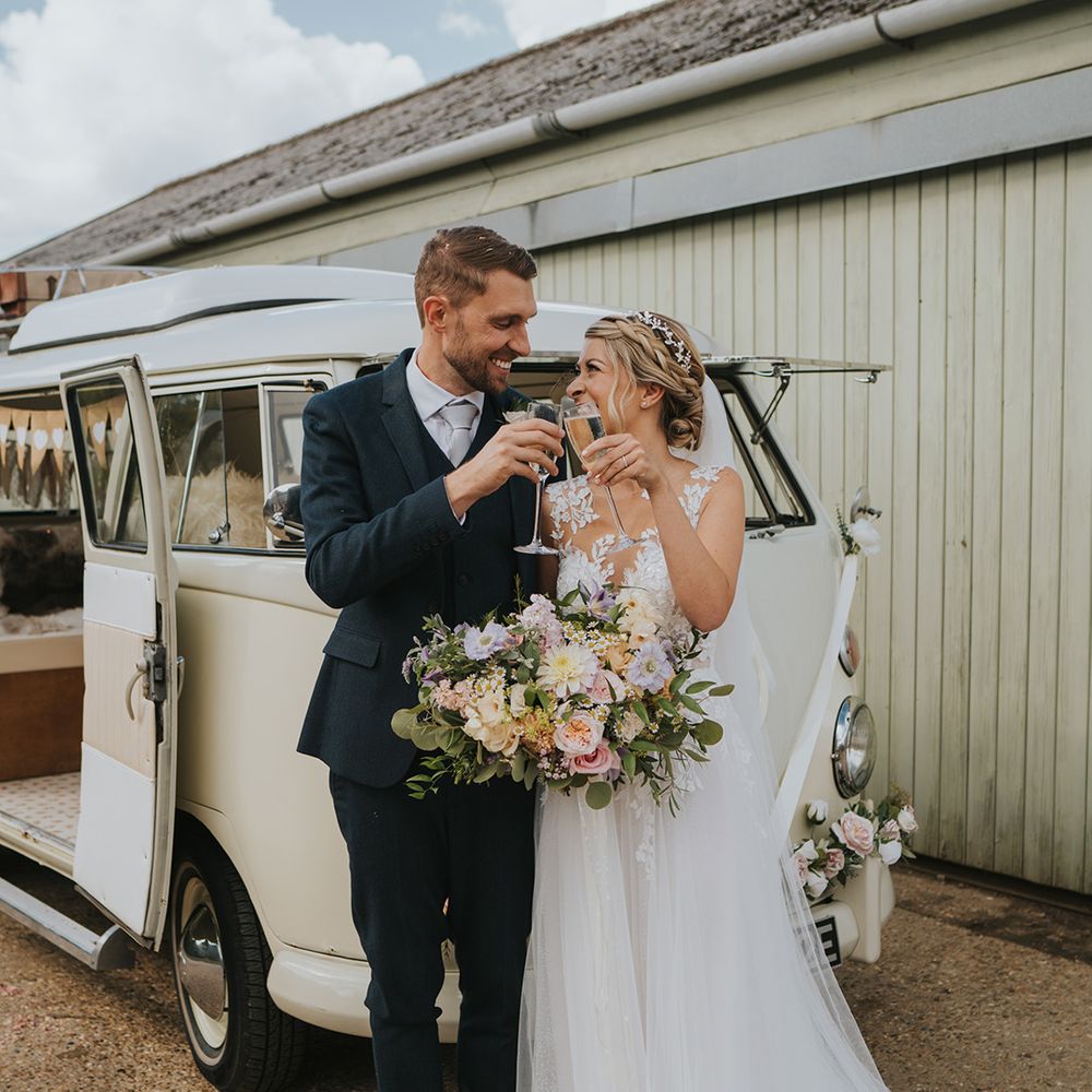 The bride and groom cheers their glasses of champagne in front of their cream VW camper van transport 