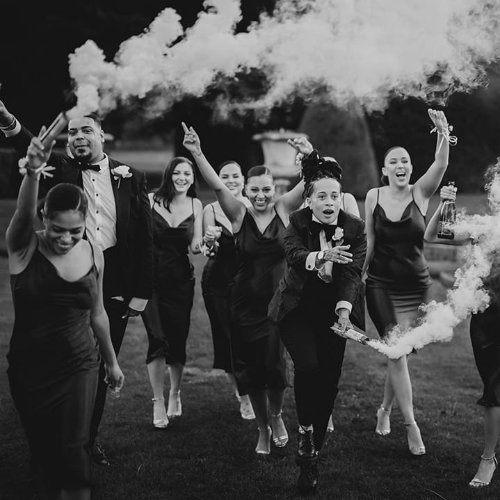 Wedding party in strappy satin midi length black bridesmaid dresses and classic black tuxedos with white rose boutonnieres doing smoke bomb wedding photos at Grittleton House wedding venue 