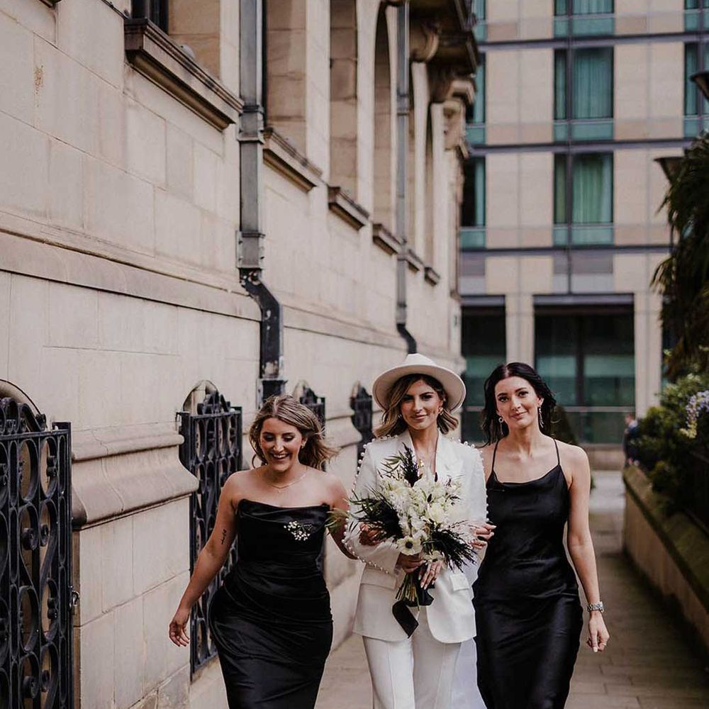 Bride in white bridal suit with fedora hat walking with bridesmaids in strappy black bridesmaid dresses 