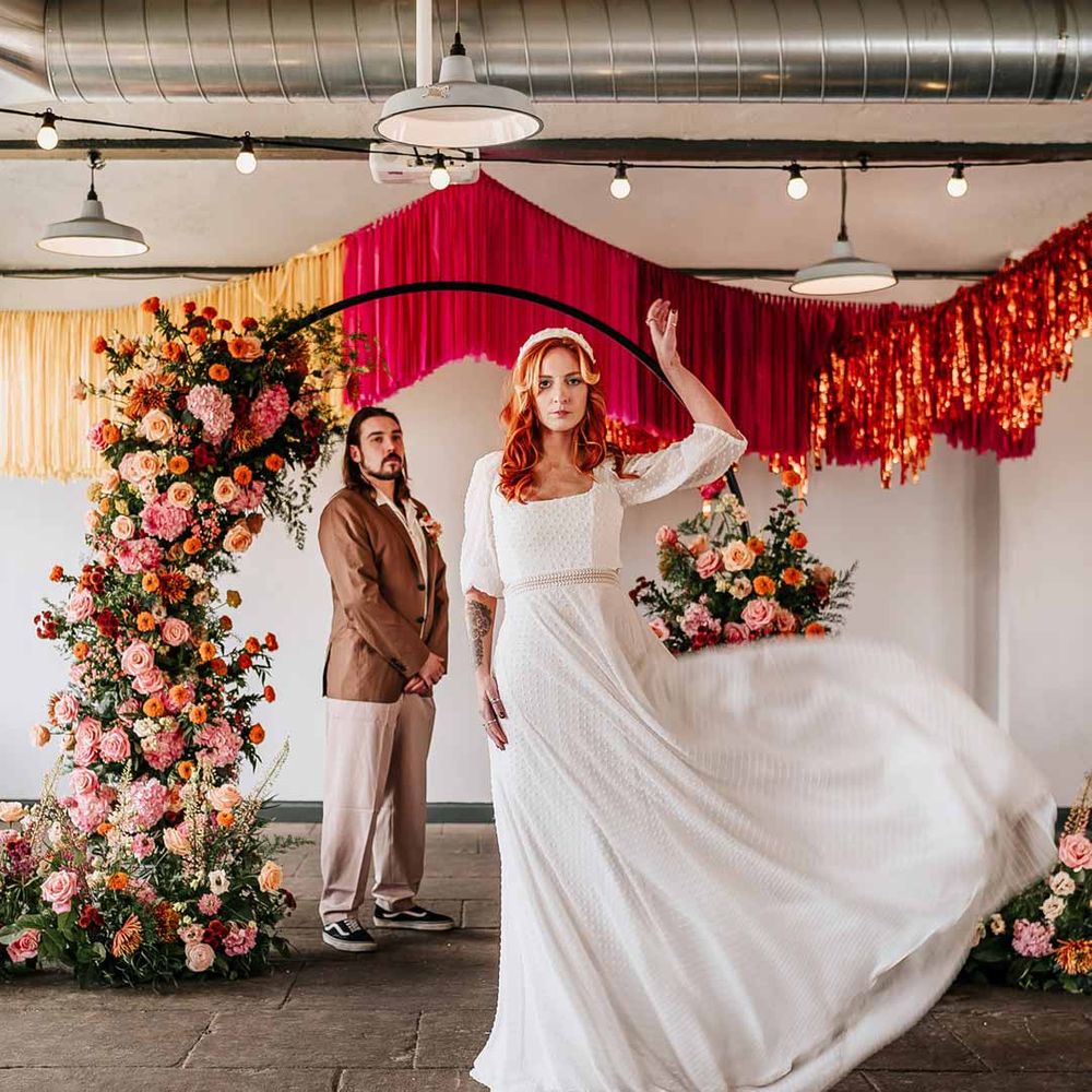 Bride in three quarter length puff sheer sleeve wedding dress with delicate polka dot overlay and square neck and white 3D applique polka dot headband with groom watching on wearing brown blazer, white shirt, grey suit trousers and vans shoes standing underneath circular floral arch with vibrant flowers and foliage attached and pink, red, yellow and metallic wedding streamers behind