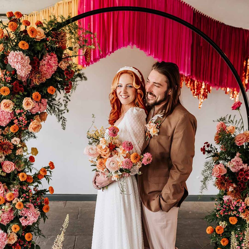Bride in three quarter length puff sheer sleeve wedding dress with delicate polka dot overlay and square neck and white 3D applique polka dot headband posing with groom in brown blazer, white shirt, grey suit trousers and vans shoes underneath circular floral arch with vibrant flowers and foliage attached and pink, red, yellow and metallic wedding streamers behind at Northern Monk Refectory Leeds