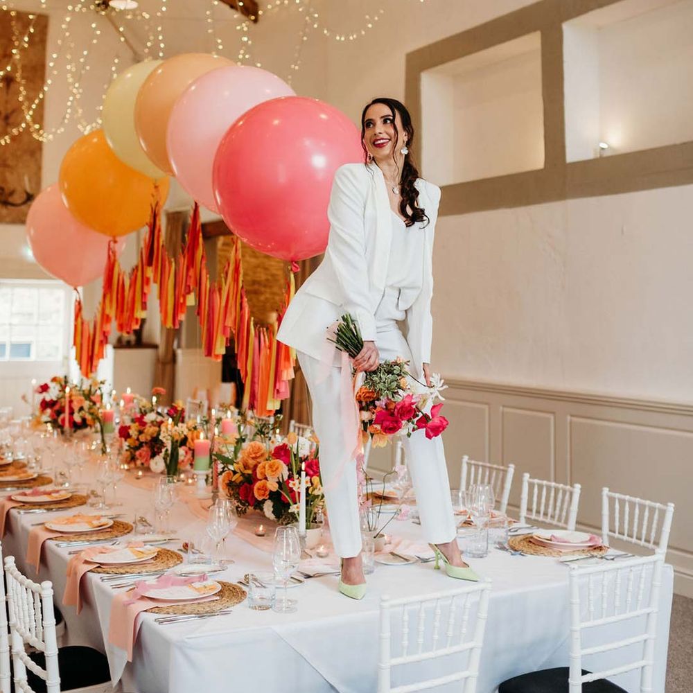 Bride in white bridal suit with silver bridal jewellery with closed toe bright green wedding heels standing on wedding tablescape at Merriscourt wedding venue with wedding tablescape with white wedding tablecloth, burlap place mats, tapered candles, pink and orange wedding stationery, and floral wedding table centrepieces with pink and orange garden roses, orange floribunda, yellow ranunculus flowers, foliage and Iceland poppies with red, pink orange and yellow balloons, wedding streamers and fairy lights at Merriscourt wedding venue