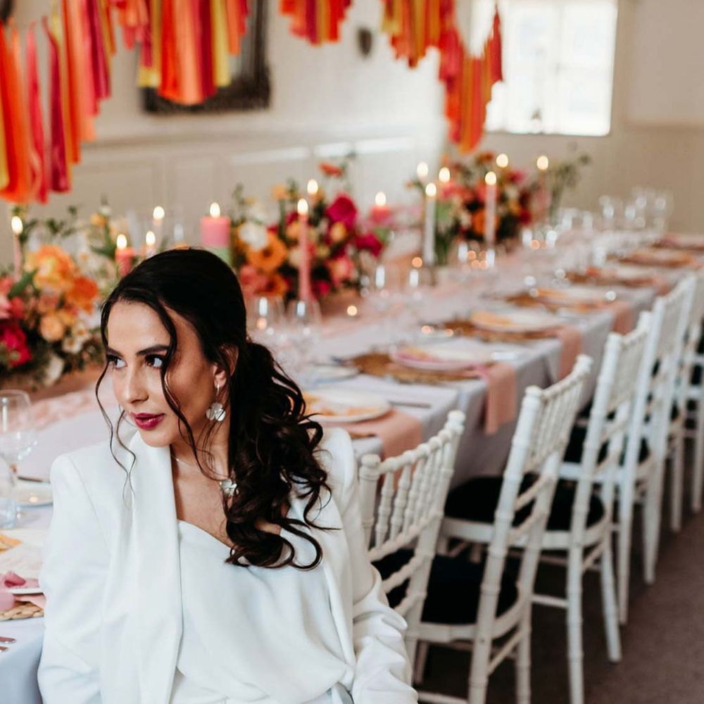 Bride in white bridal suit with silver bridal jewellery sitting in by wedding tablescape with white wedding tablecloth, burlap place mats, tapered candles, pink and orange wedding stationery, and floral wedding table centrepieces with pink and orange garden roses, orange floribunda, yellow ranunculus flowers, foliage and Iceland poppies at Merriscourt wedding venue