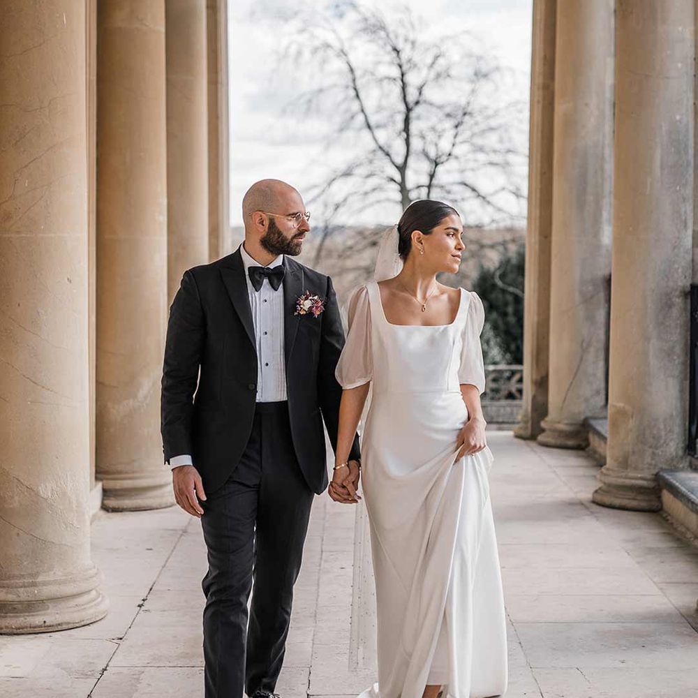 Groom in classic black tuxedo with bowtie and wildflower boutonniere walking with bride in short sheer puff sleeve wedding dress with square neck and wrist loop with sheer pearl embellished bridal veil at Buxted Park
