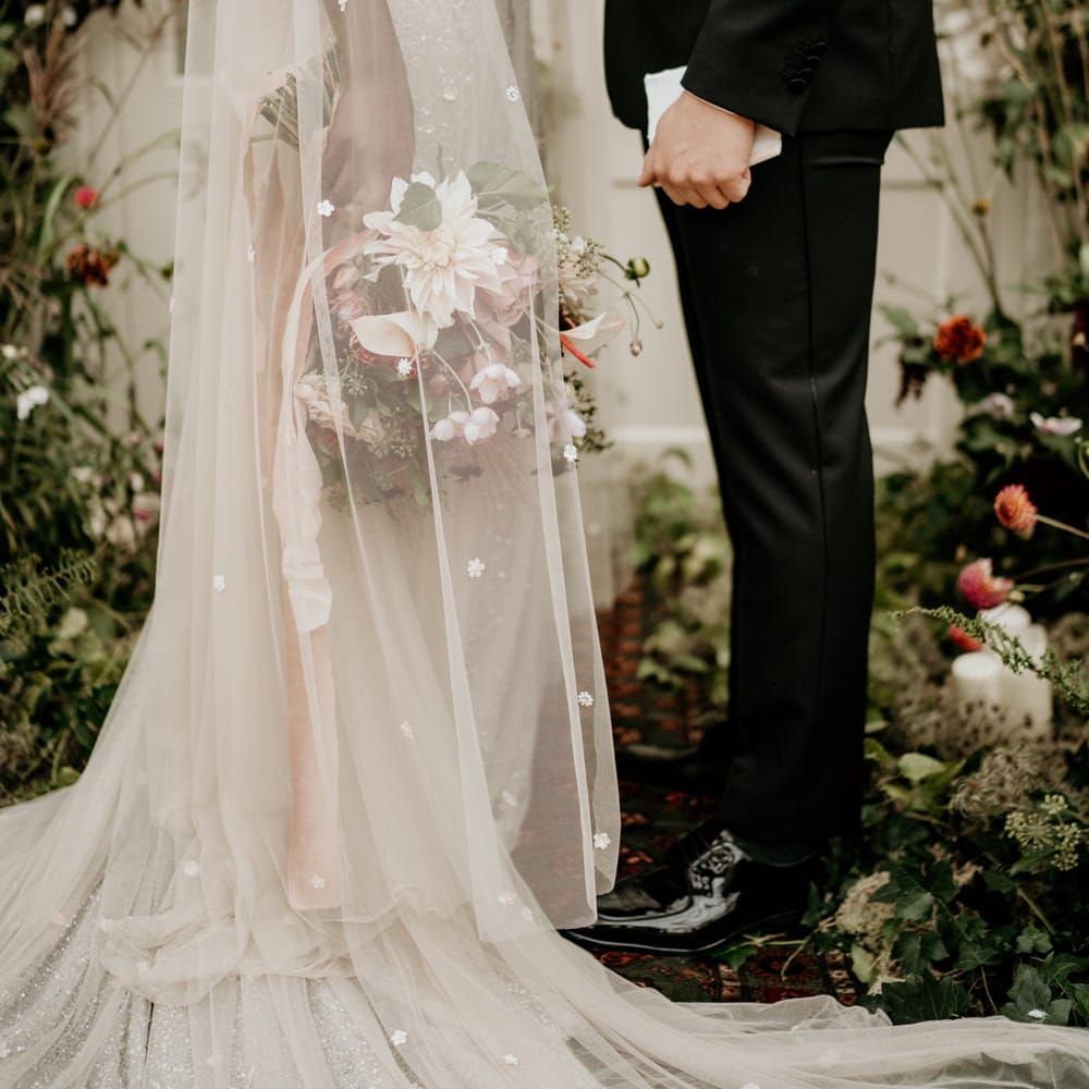 Bride in a Julita Bridal London wedding dress with embellished detail and cape standing at the altar with her groom in a black tuxedo 