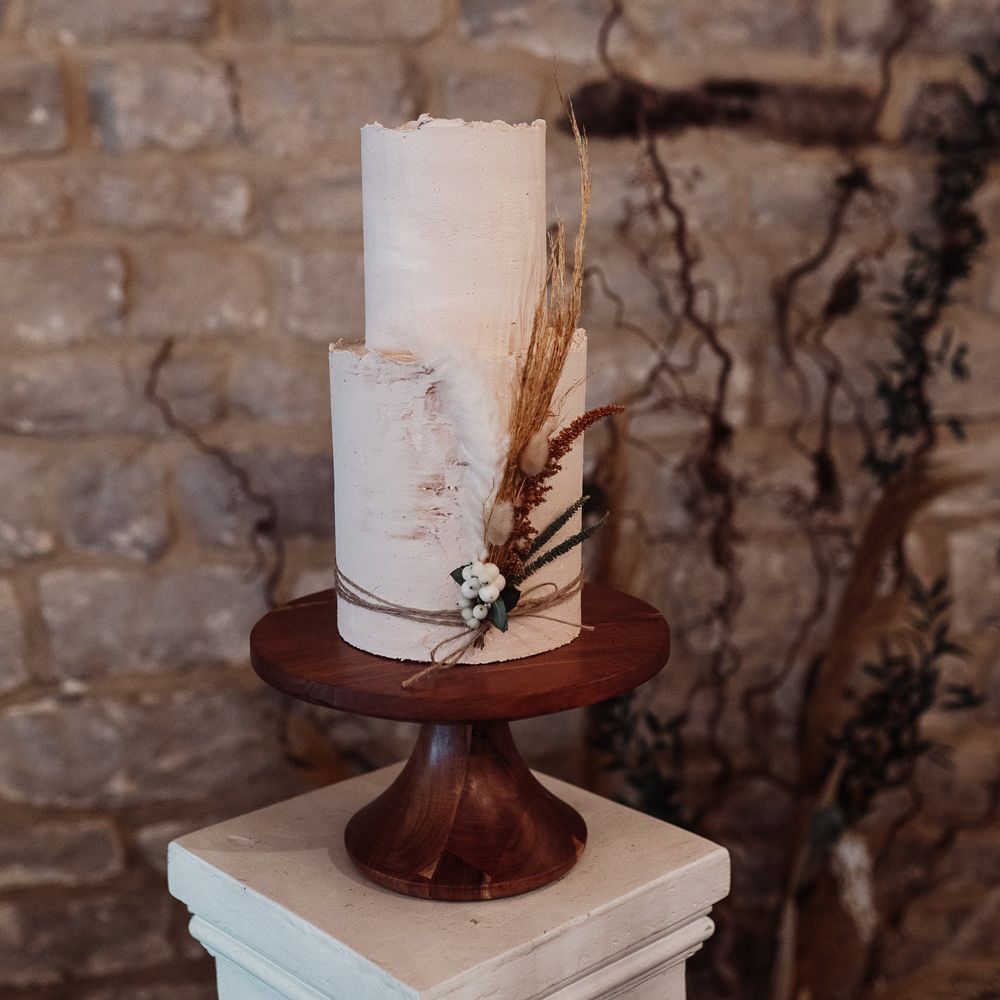 Tall two tier wedding cake on a wooden cake stand with dried grass tied around the cake for decoration
