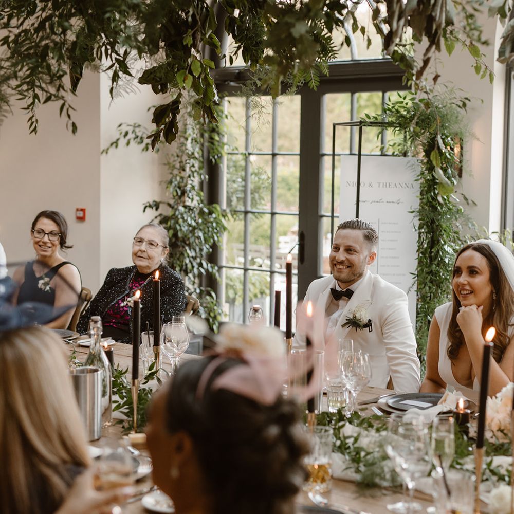Groom in white tuxedo jacket, bride in Made With Love wedding dress and veil and guests sit at table listening to speeches in the Fig House decorated with greenery