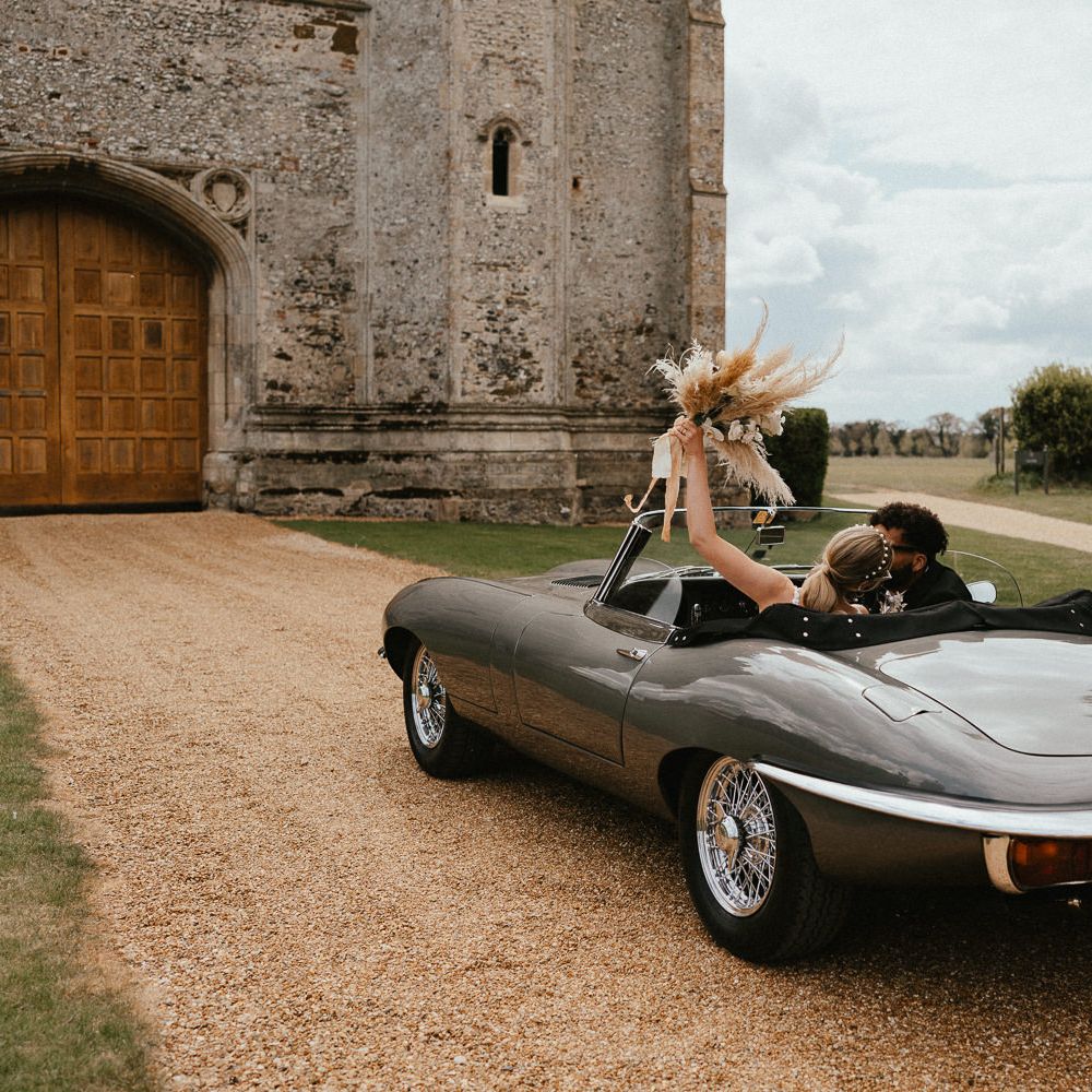 Bride and groom kissing in a vintage wedding car with the bride holding her dried flower bouquet in the air 